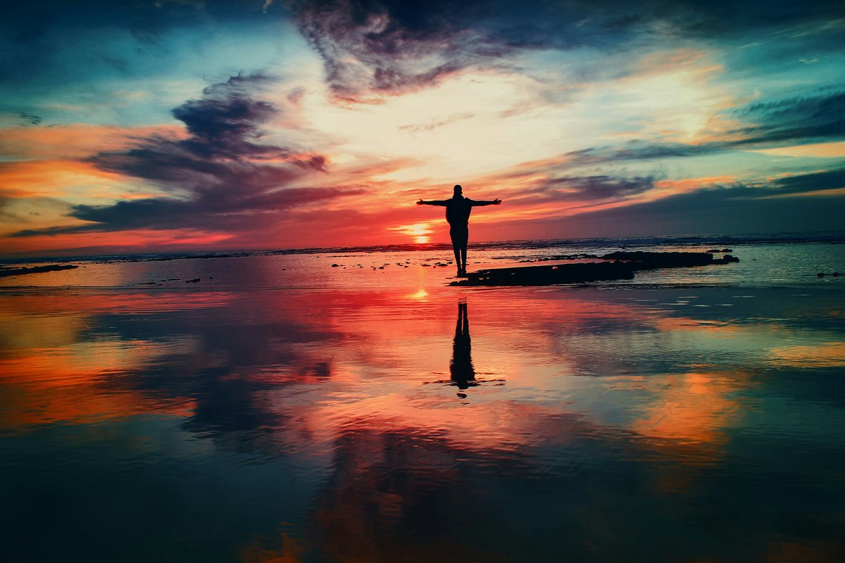 A person standing with arms outstretched at the water's edge at sunset